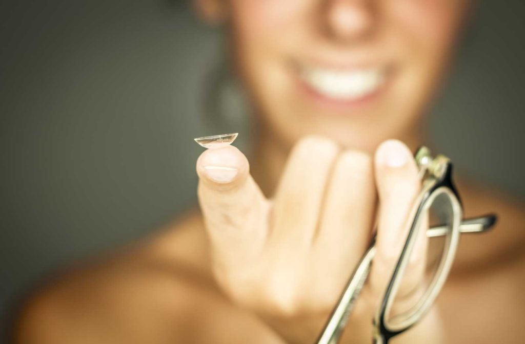 smiling woman out of focus holding up choice of contact lens and glasses
